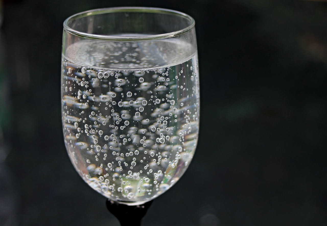 Bicchiere trasparente di acqua frizzante con bolle visibili, limone fresco accanto e superficie di marmo bianco