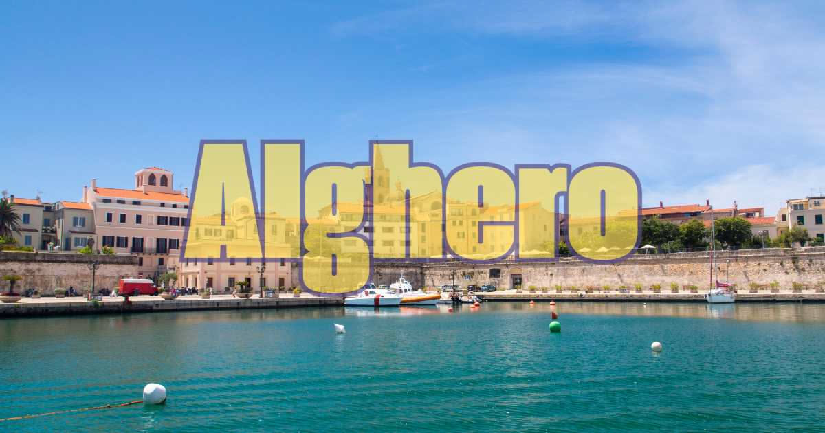 Veduta aerea del centro storico di Alghero con mura catalane, porto turistico, case dai tetti rossi e spiaggia sabbiosa, con mare blu sullo sfondo