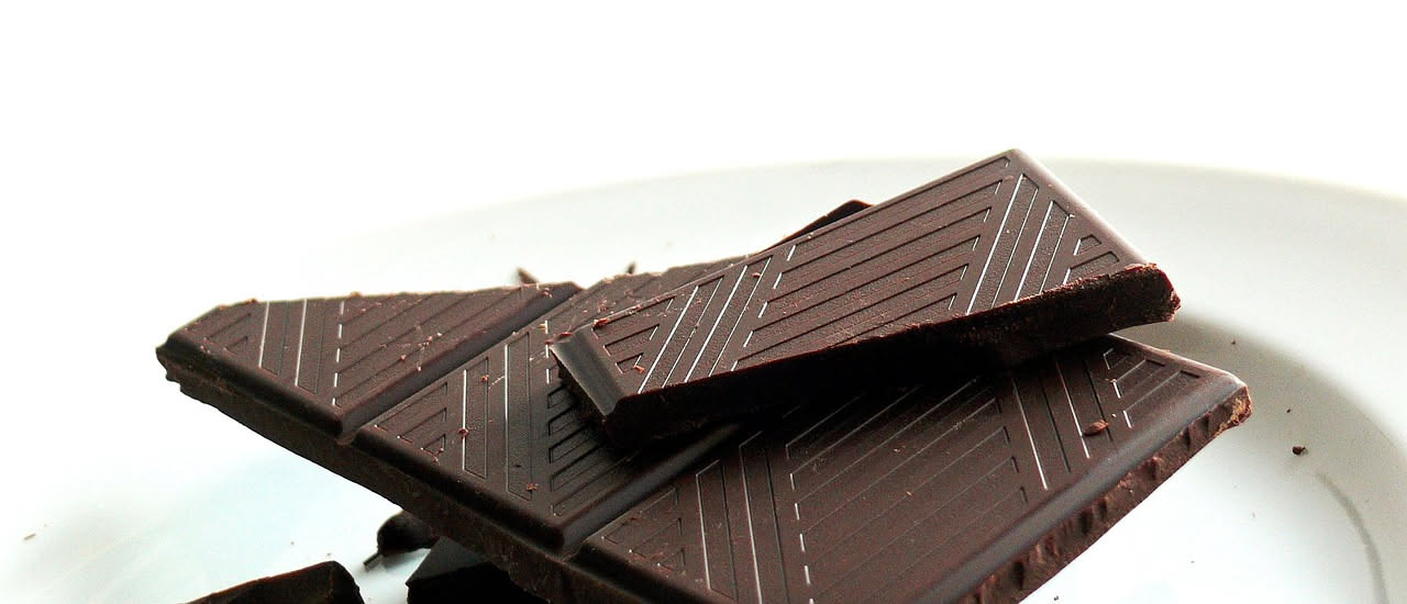 Cioccolata fondente spezzata in quadrotti su un piatto bianco, con accanto cacao in polvere e fave di cacao, su uno sfondo di legno chiaro