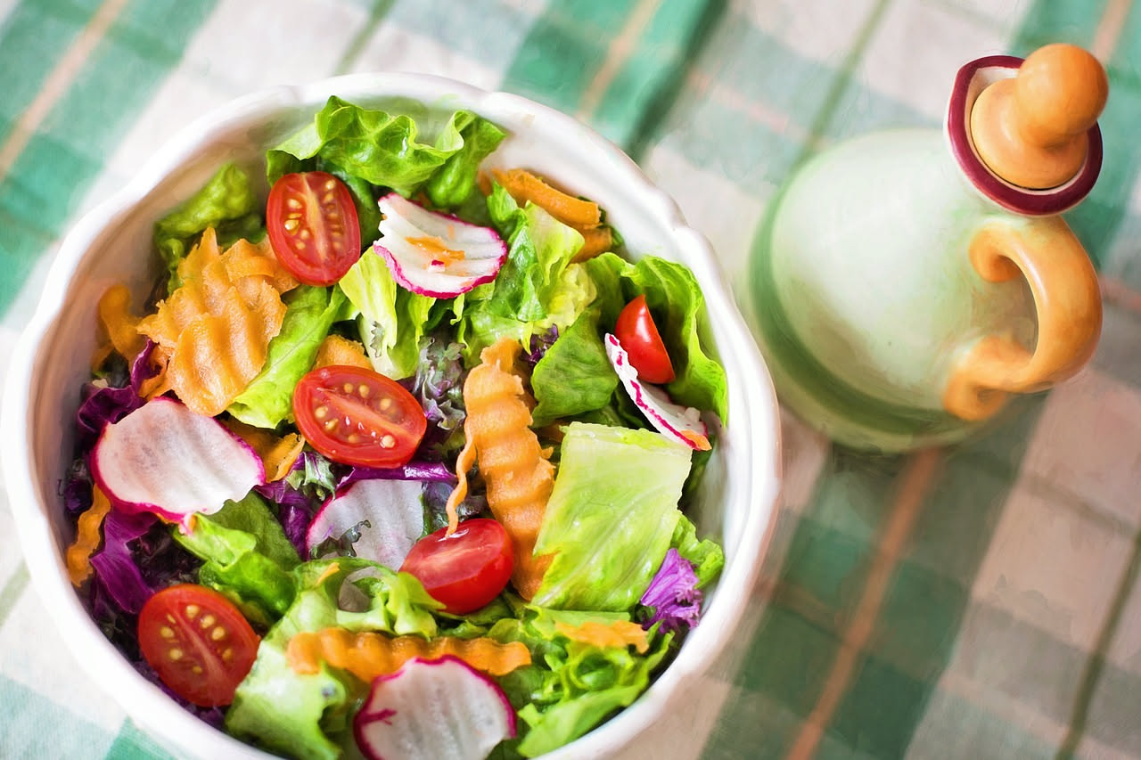 Piatto di insalata mista con verdure fresche di colore verde e rosso su un tavolo bianco, accanto a un piatto con pasta e pollo in preparazione