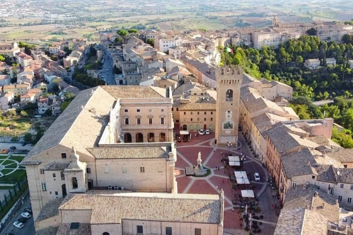 Panorama di Recanati con le colline marchigiane sullo sfondo, piazza centrale con edifici rinascimentali in mattoni rossi e cielo azzurro