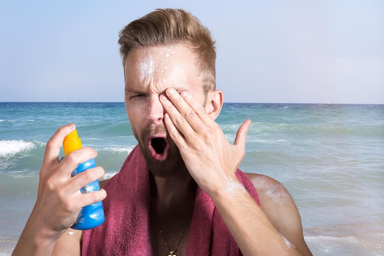 Persona in spiaggia durante una giornata soleggiata estiva, con gocce di sudore visibili sulla fronte e sulla pelle, con il mare sullo sfondo