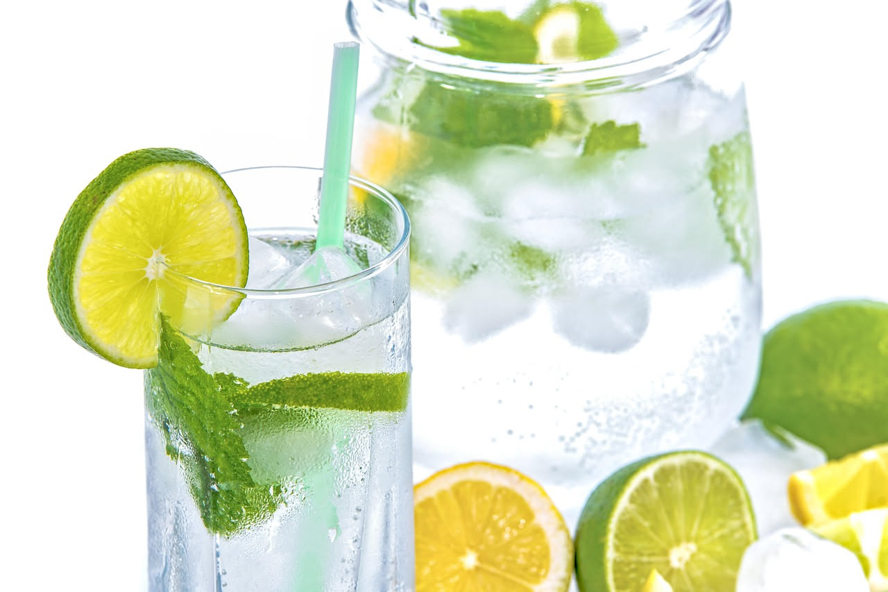 Bicchiere trasparente pieno di acqua fredda con ghiaccio e fette di limone, su sfondo bianco con gocce di condensa, durante giorno soleggiato